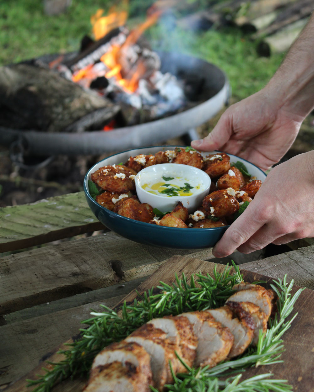 Hand placing a dish onto a wooden table with a fire pit behind